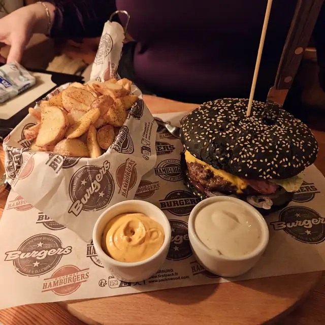 Burger z dzika w czarnej bułce na desce, z ćwiartkami ziemniaków i sosami
