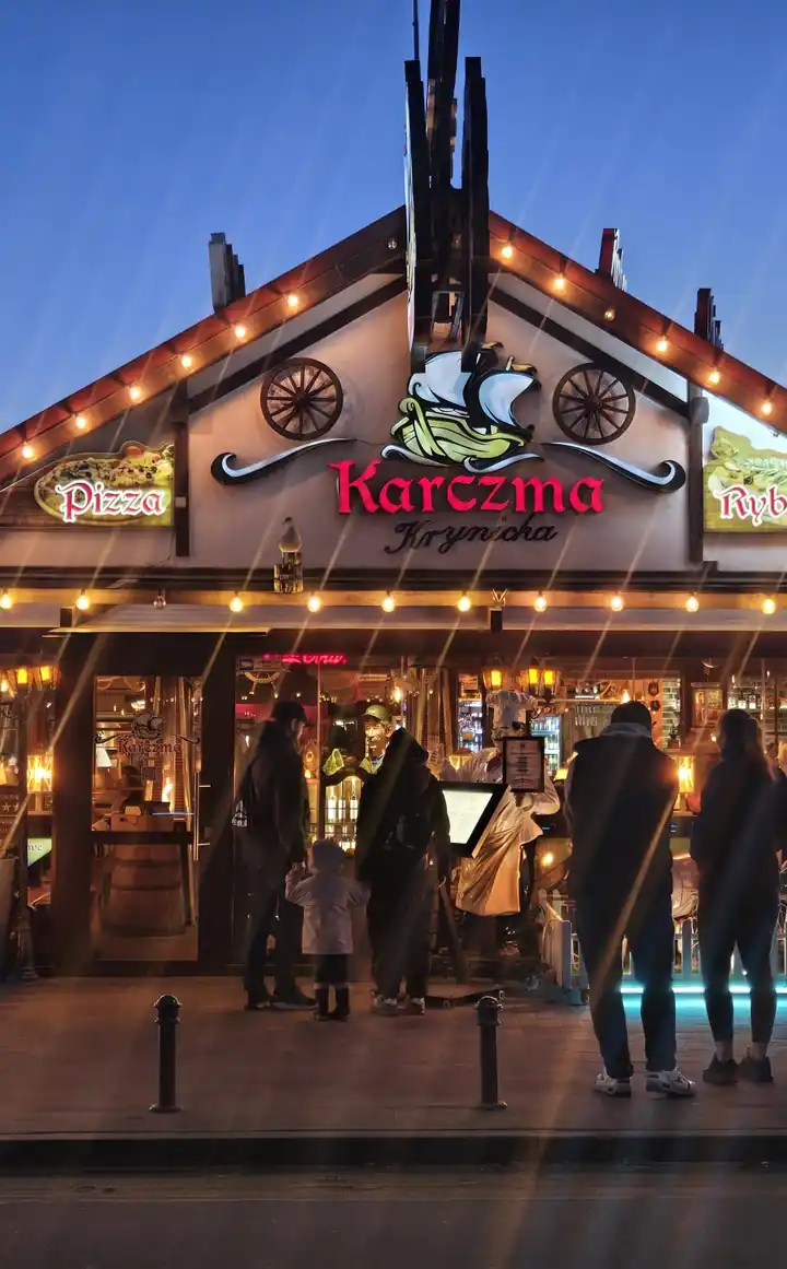 Karczma Krynicka o zmierzchu: drewniana fasada z czerwonym napisem Karczma obwieszona girlandami świateł, przed wejściem goście