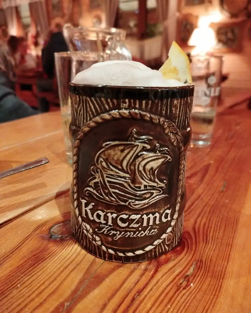 Ceramiczny kufel z logo Karczmy Krynickiej i pianą, na stole w ciepłym świetle